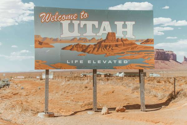 A grey-blue sky and orange landscape matches the colors of a sign reading "Welcome to Utah: Life Elevated"; the sign also bears a grey-blue background and the orange terrain of Arches National Park