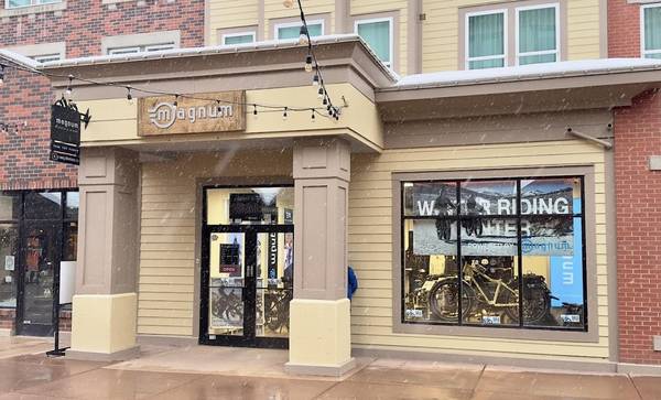 Magnum Bikes Park City store exterior in brown and yellow neutrals with partial brick exterior and a wooden logo sign; snow is falling