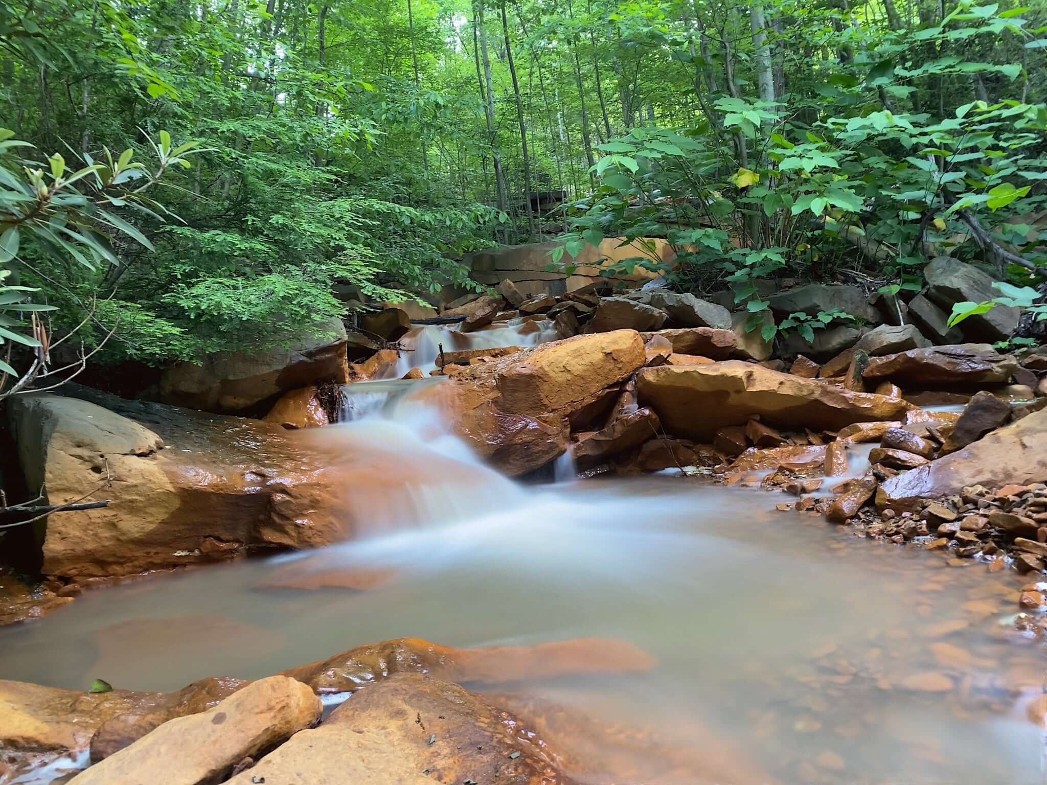 A long exposure photograph creates a silky effect on a small waterfall over brown rocks, surrounded by lush greenery