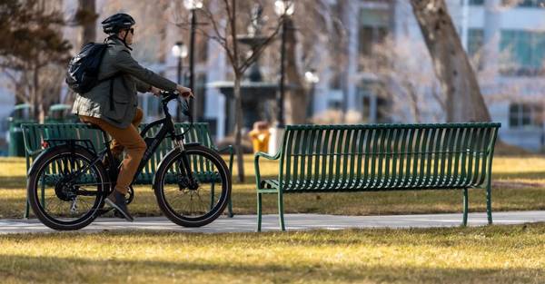 Rider in black helmet, khaki jacket, orange pants, and sneakers rides a Magnum Metro 750 electric bike through a park, on a paved trail between two benches