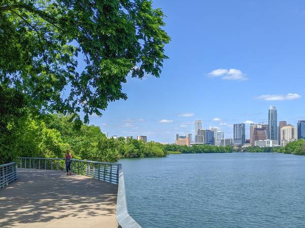 Sunny view of Lady Bird Lake in Austin Texas along a boardwalk section of the Ann and Roy Butler Hike and Bike Trail; a jogger runs near the railing on the boardwalk