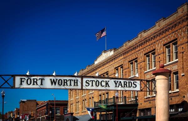Vintage sign spanning 2 pillars across the road, reading "Fort Worth Stock Yards," in front of old brick buildings and a clear blue sky