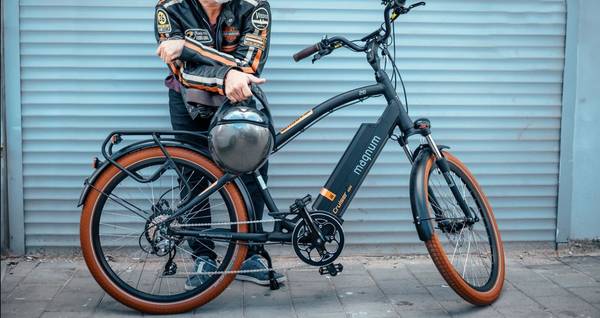 Rider in black and orange motorcycle jacket carries a helmet and leans on their Magnum Cruiser 2.0 black and orange e-bike in front of a light blue garage door