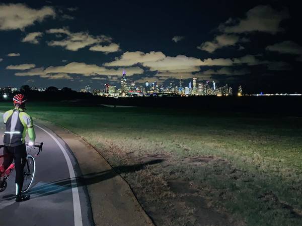A paved bike path with an illuminated bike rider shining a light on a green lawn; in the distance, the full city skyline of Chicago is illuminated, and white clouds can be seen reflecting the city lights