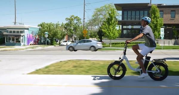 A rider in a blue helmet, grey t-shirt, and white shorts rides a white Magnum Premium 3 Low-Step Folding e-bike on a bike path adjacent to a road; we see glimpses of green grass and trees as well as buildings across the street