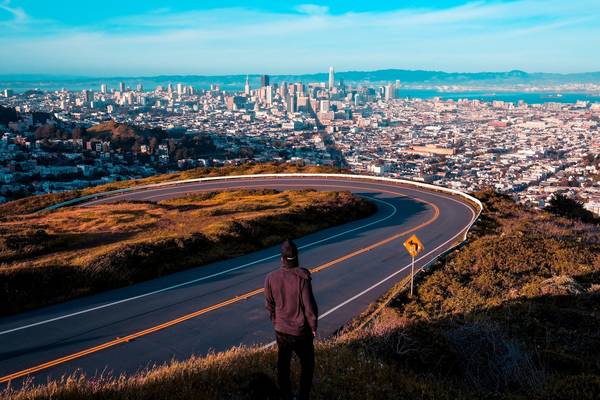 A person in casual black clothing faces away from the camera looking at the view from Twin Peaks in San Francisco; the full city skyline and the bay can be seen in the distance
