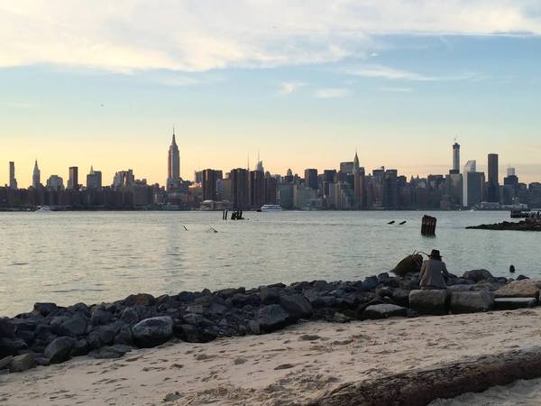A person in a wide brimmed hat sits on a rock on the beach in Brooklyn, overlooking the water toward Manhattan at sunset