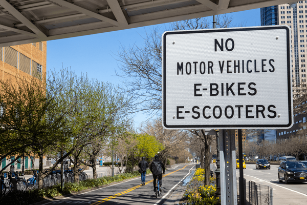 A paved bike path runs underneath a bridge next to a sign reading "No motor vehicles e-bikes e-scooters"