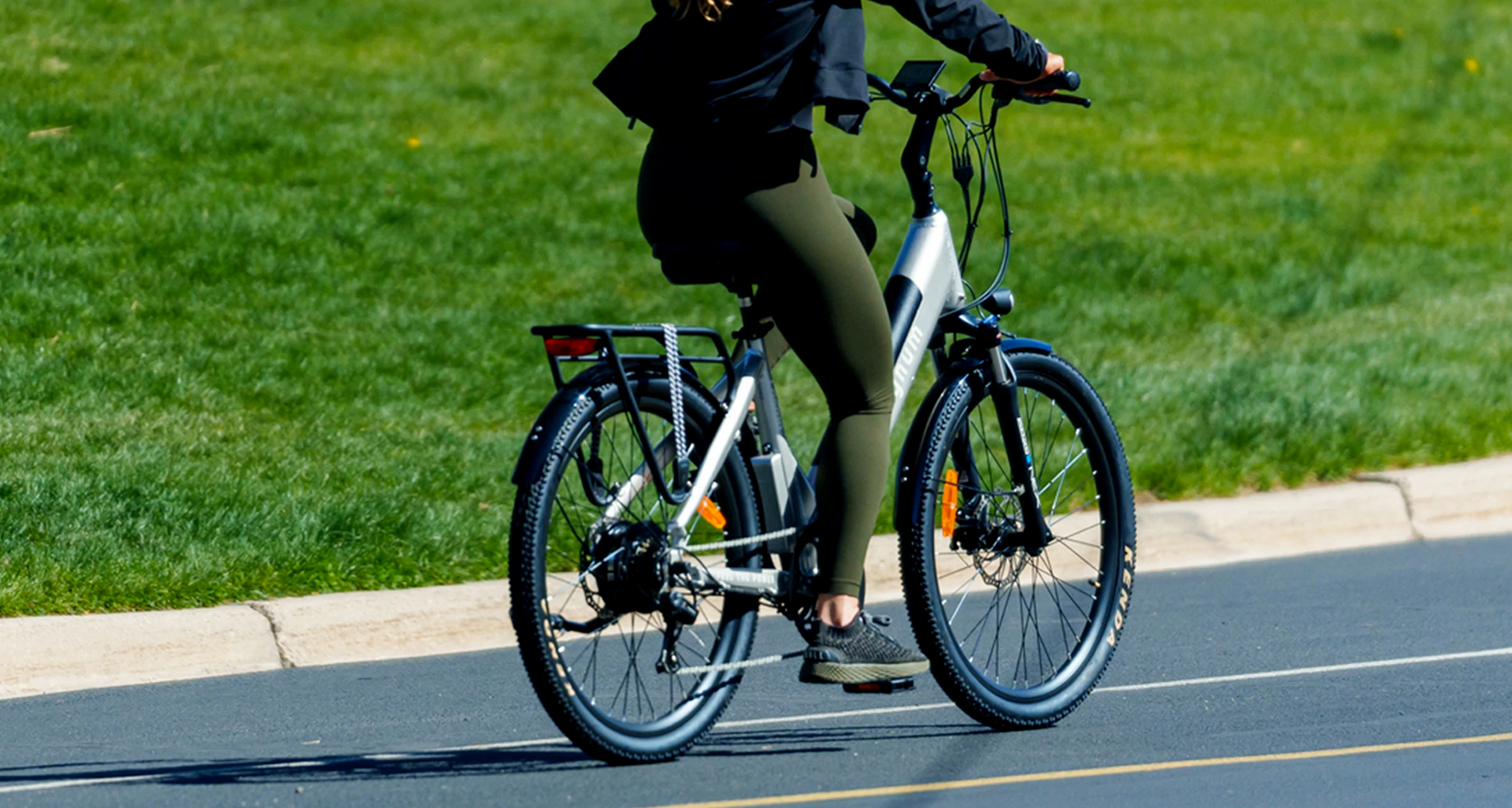 Rider with olive green leggings and black sneakers on a Silver Magnum Cosmo e-bike in the street in front of a stretch of green grass
