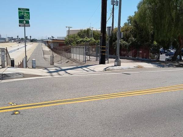 A green road sign with a picture of a bicycle indicates the start of the Coyote Creek Bikeway for e-bikes along a paved road in an industrial area