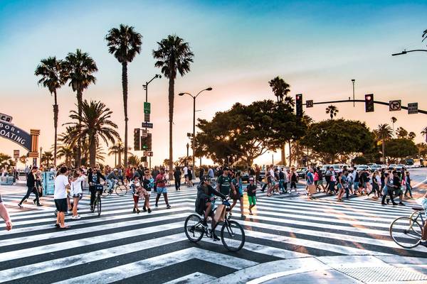 Bicyclists and pedestrians navigate a busy striped crosswalk on Santa Monica Boulevard at sunset amid tall palm trees and traffic signals