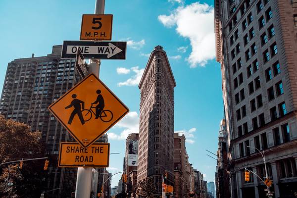 A yellow diamond sign depicts a pedestrian and e-bike rider above a text sign reading 'Share the Road', displayed on a post in Manhattan with the Flatiron Building in full view