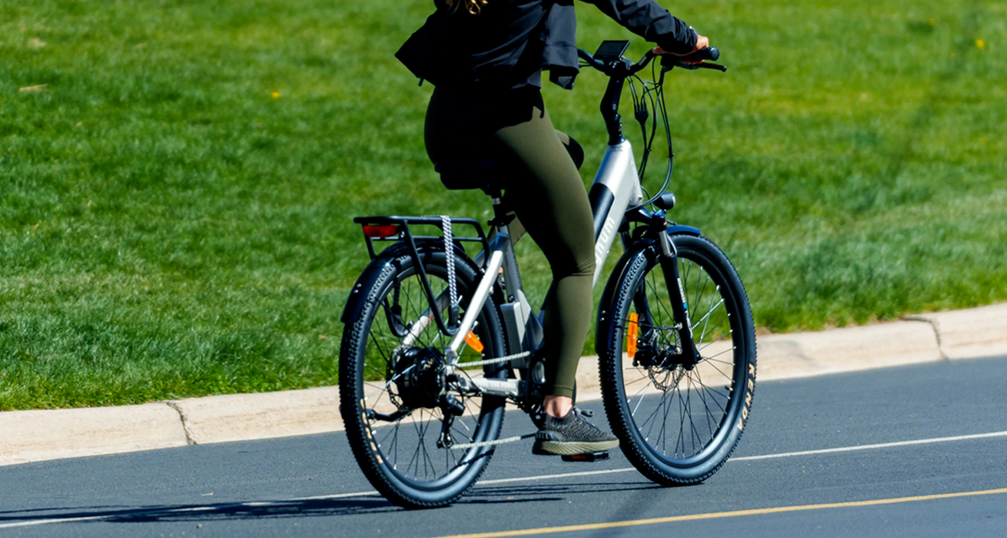 Rider with olive green leggings and black sneakers on a Silver Magnum Cosmo e-bike in the street in front of a stretch of green grass