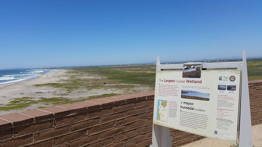 A clear cloudless day with ocean waves, sandy beach, and green ocean plant life on the beach. A sign on a dirt pathway with brick guardrail displays both Spanish and English text and reads "The Largest Coastal Wetland"