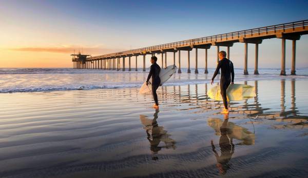 Two surfers in black bodysuits walk across the sand carrying their boards toward the waves for a sunset surf near a pier in San Diego