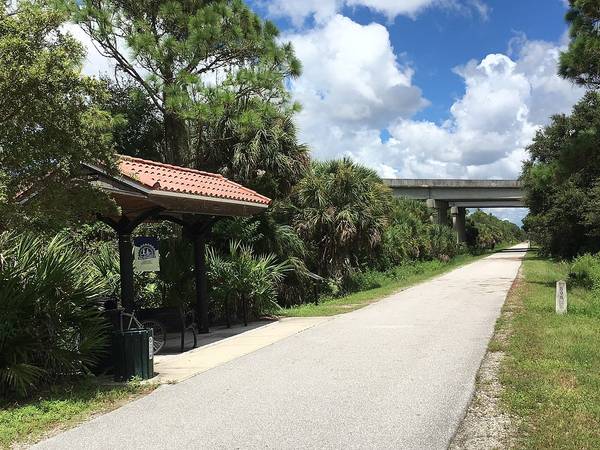 A shelter with bench along Sarasota's green and e-bike friendly Legacy Trail leading under a road beneath blue sky with white clouds