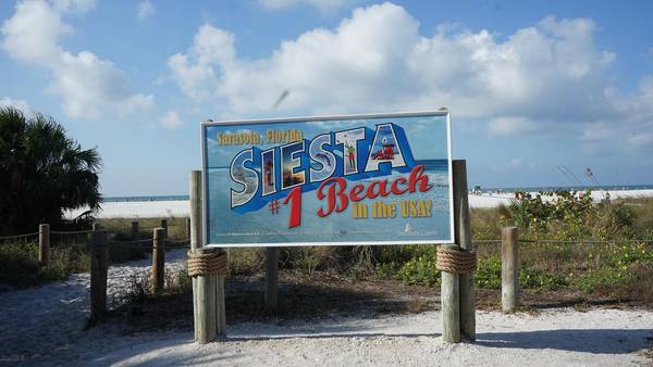 White sand in the foreground and a path leading down to the beach, with a colorful blue, red, and yellow sign reading "Sarasota, Florida — Siesta — #1 Beach in the USA!"
