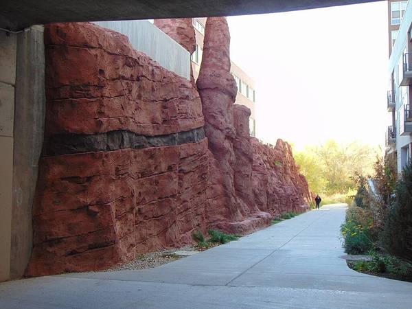 An underpass on Parley's Trail in Salt Lake City runs between a red rock wall and a residential building; a pedestrian walks the paved multi-use trail in the distance