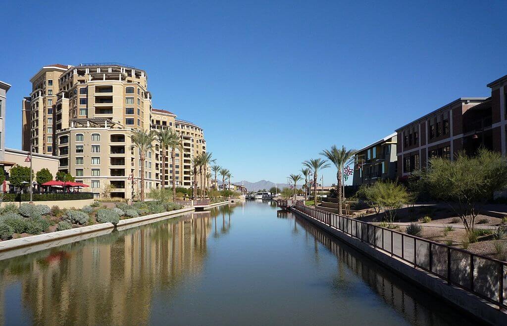 View of the Arizona Canal at the Scottsdale Waterfront with tall buildings, fountains, and palm trees in the foreground and a blue sky with white clouds in the background