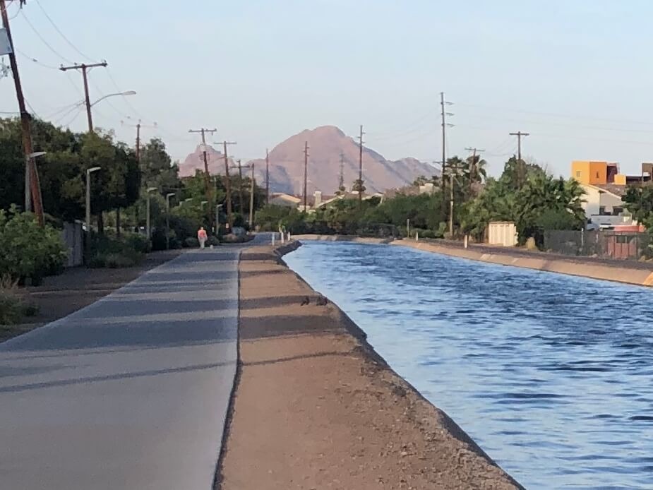The Grand Canal Path in Phoenix, Arizona. The paved trail stretches into the distance, flanked by lush greenery on either side. The path is empty except for a lone pedestrian in the distance. The sky is clear and blue above.