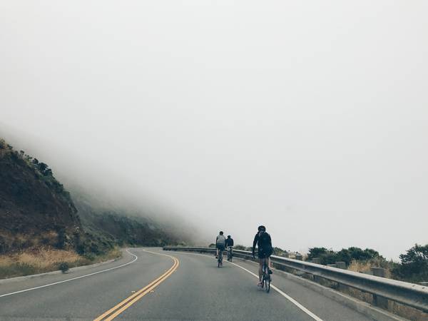 3 bicyclists riding down a winding empty two-lane road surrounded by fog and hills