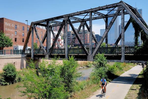 A cyclist rides on a paved e-bike path alongside a canal that flows under a bridge in Denver Colorado