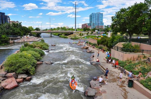 A view of South Platte River Trail in Denver, with a busy paved path filled with visitors stretching through green grass, trees, and bushes. The trail runs alongside a river, where a kayaker floats, and a bridge is visible in the background.