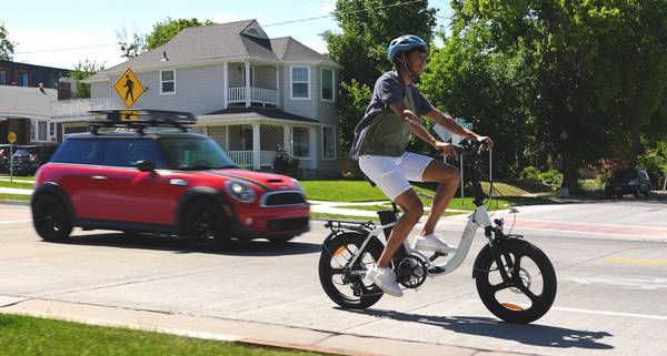 A rider cruises in the bike lane on a white Magnum Premium 3 Low-Step e-bike as a small red 2-door car prepares to pass by. The rider wears a blue helmet, green and gray t-shirt, white shorts, and white sneakers
