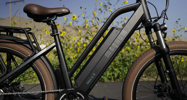 A black Magnum Metro S electric bike close-up, with only part of each wheel visible and the stem out of frame. Yellow wildflowers grow tall in the background