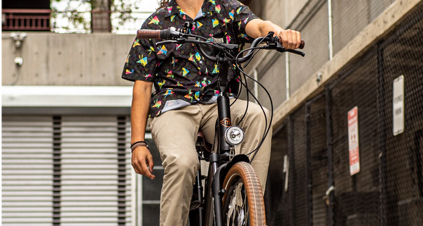 A rider shown from the neck down wears a black shirt with neon geometric patterns and khaki pants, riding a Magnum Low Rider 2.0 electric bike in a relaxed pose with one hand on the handlebars and the other hanging at their side