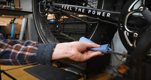 A hand holds a blue cloth between thumb and index finger, wrapped around a Magnum e-bike chain