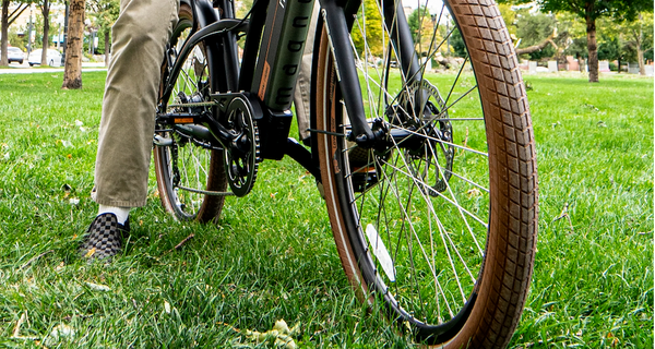 Angled front view of a black Magnum Low Rider e-bike with brown tires; a rider wears checkered slip-on shoes and khaki green pants, resting one foot on the ground and one on a pedal