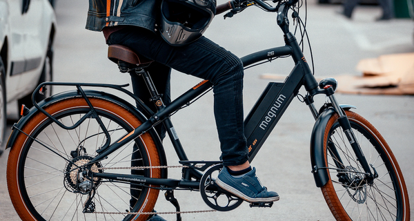 A rider wearing a black and orange leather jacket, black pants, and blue sneakers straddles the Magnum Cruiser e-bike with one foot on a pedal; the quick release lever on the front wheel is clearly visible since the wheel is angled away from the camera