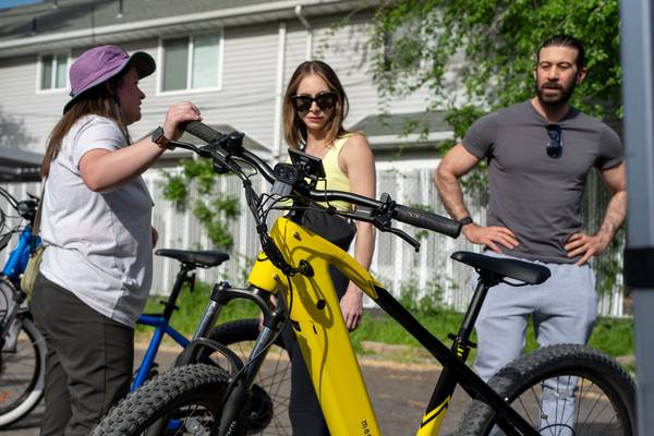A woman white t-shirt and pink sunhat shows off a yellow Magnum Vertex e-bike to a woman in sunglasses and man in gray t-shirt