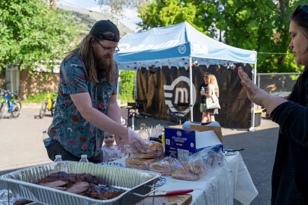 A man in turquoise and pink floral shirt grabs burger buns from a bag to serve a guest, while a Magnum Bikes tent can be seen in the background