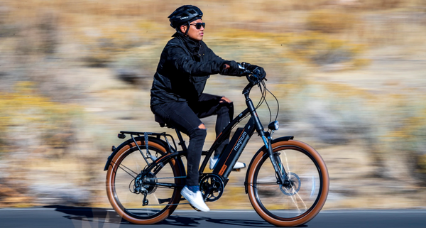 A rider in black helmet and sunglasses, dressed in all black with white slip-on sneakers, rides a black and orange Magnum Low Rider 2.0; the camera focuses on the rider in motion, so the background is blurred