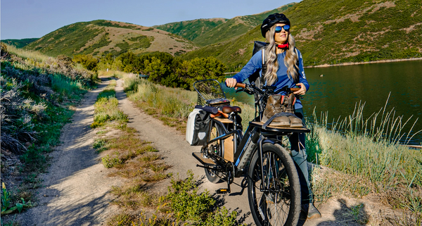 A rider with long blonde hair wears a helmet and sunglasses as she walks her fully loaded Magnum Payload cargo e-bike along a dirt path. The e-bike carries hiking boots, camping gear, and more.