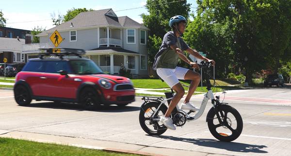 A rider in helmet, green shirt, white shorts, and white sneakers rides a white Magnum Premium 3 folding e-bike in a bike lane as a small red car approaches from behind.