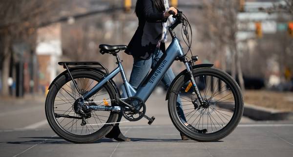 A rider in black blazer, denim pants, and black angle boots walks a blue Magnum Cosmo X e-bike across the street