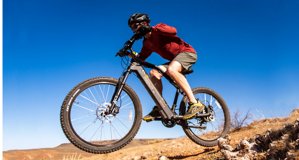 A man in red long-sleeve shirt and grey shorts rides a Magnum Summit 27.5" mountain e-bike over a jump on rocky terrain, with cloudless blue sky behind him.