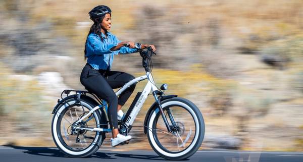 A woman in a denim jacket and black pants rides a white Magnum Ranger e-bike. The background is blurred to show fast movement.