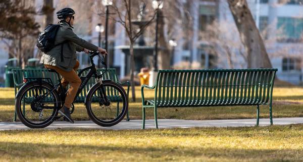 A man in sunglasses, gray jacket, and khakis rides a Magnum Metro 750 e-bike through a park; he's on a paved path running between two green benches, with trees and grass surrounding.