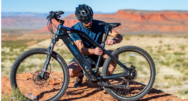 A rider in black helmet and blue t-shirt stops to adjust the height of his seatpost on his black and blue Magnum Summit e-bike with a beautiful desert backdrop