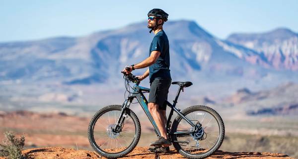 A rider in black helmet, blue t-shirt, black shorts, black sneakers pauses to take in the desert view while straddling a black and blue Magnum Summit e-bike