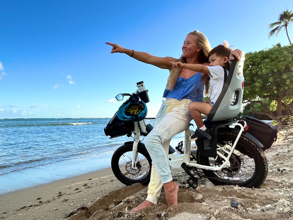 A rider in blue tank top and white joggers leans on her Magnum Pathfinder e-bike with arm around a child in child seat mounted to the e-bike. They are on a beach in the sand, and both point at something in the distance