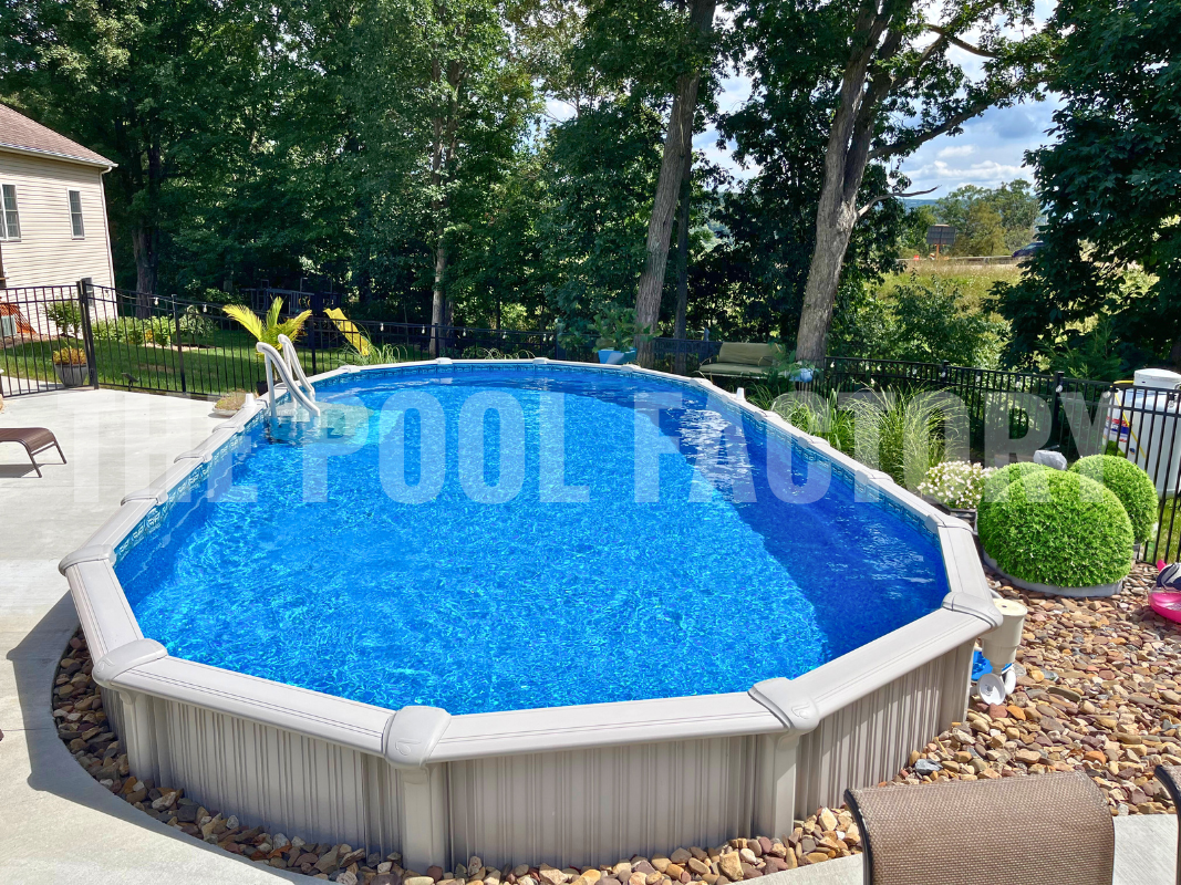 Semi-inground oval pool with concrete patio, decorative stone, and tall trees backdrop