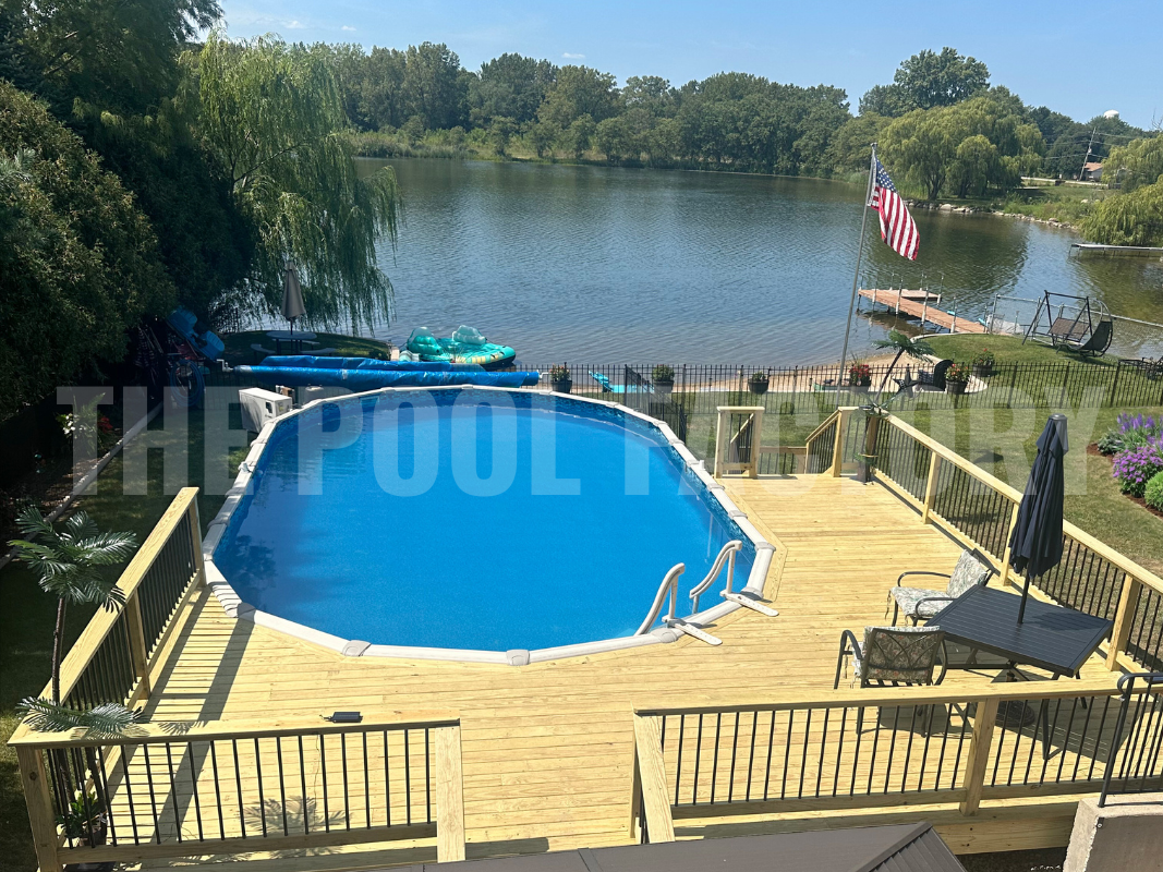 Oval above-ground pool with deck and lake view surrounded by trees