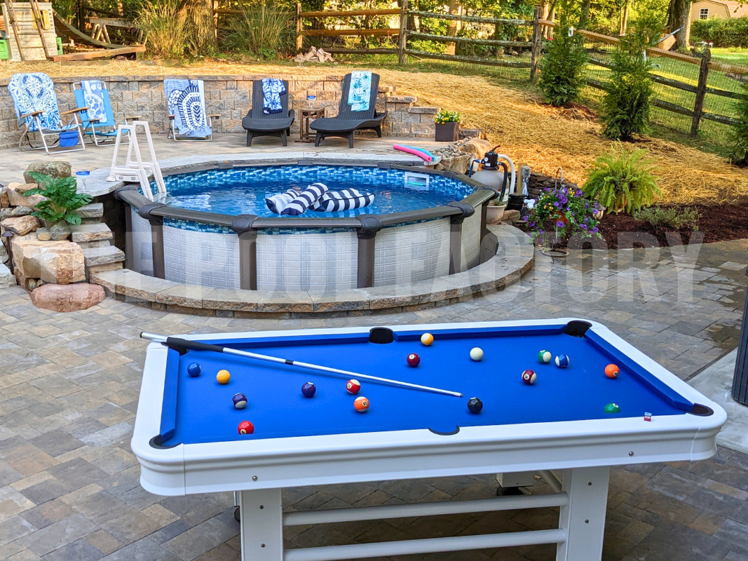 Semi-inground round swimming pool with stone patio, pool table, lounge chairs and lush landscaping