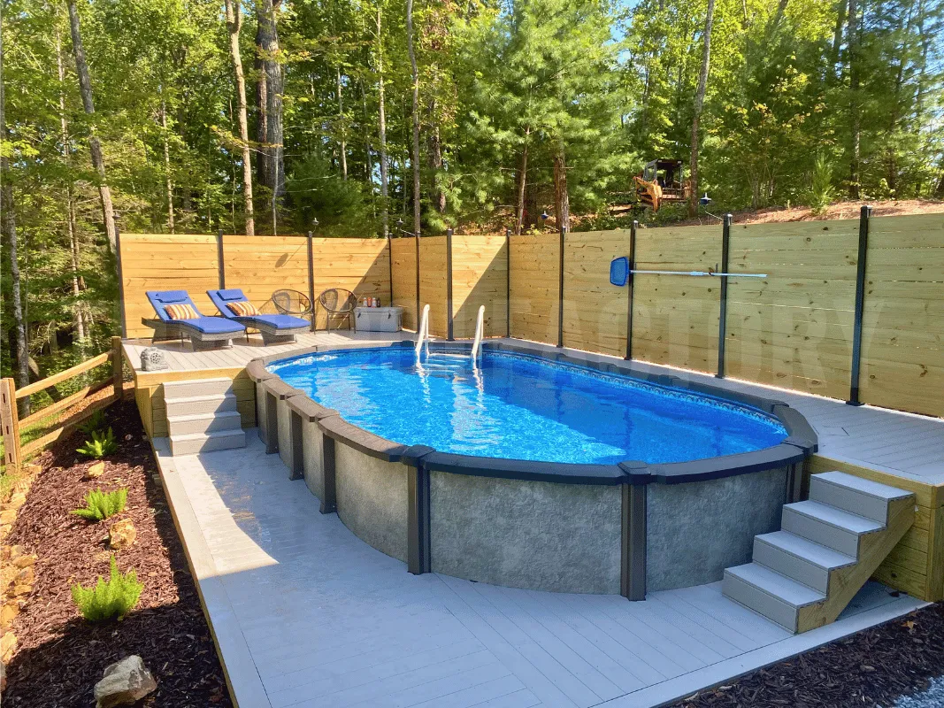 Semi-inground oval pool with concrete patio, partial wooden deck, and sun loungers under bright blue skies
