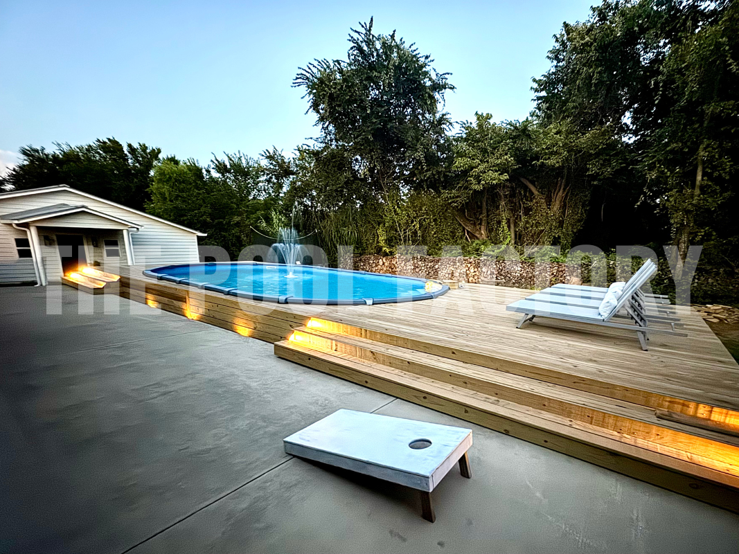 Oval semi-inground saltwater pool with night lights, surrounded by trees and full deck